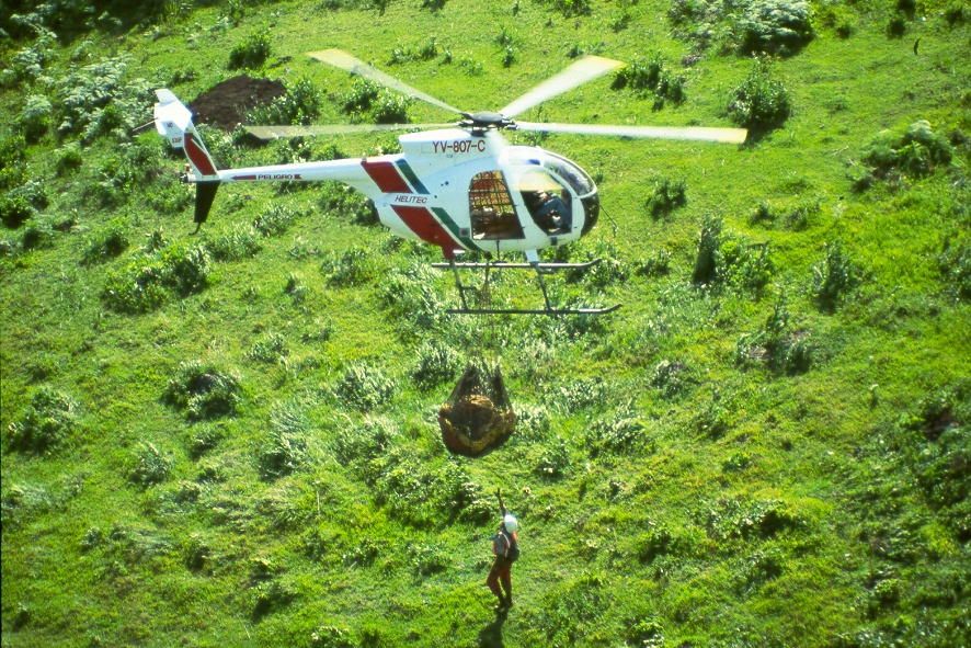 Helicóptero Helitec (YV-807-C) transportando carga con eslinga sobre pastizal, con personal de tierra
