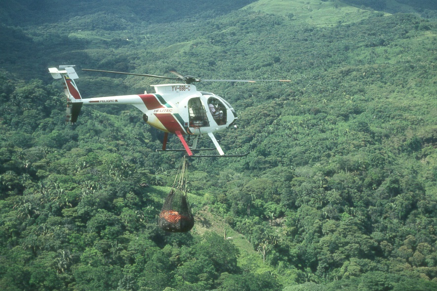 Helicóptero Helitec (YV-598-C) transportando carga con eslinga sobre terreno montañoso boscoso